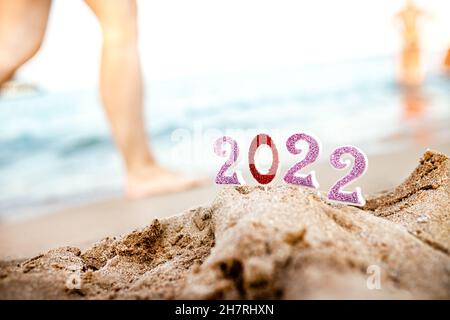 Zahlen 2022 auf Sand vor dem Hintergrund des Meeresstrandes im Resort in südlichen Ländern mit Füßen von Urlaubern, die bei Sonnenuntergang und Celebrati auf dem Wasser spazieren Stockfoto