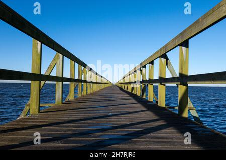 Lange, wettergetragene Pier-Anlegestelle mit Holzgeländern und Planken, die zum Meereshorizont mit klarem blauen Himmel führen Stockfoto