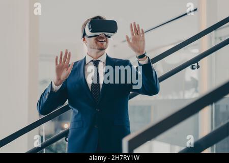 Junger, aufgeregter europäischer Geschäftsmann in formeller Kleidung, der im Büroinneren mit einem VR-Headset steht Stockfoto