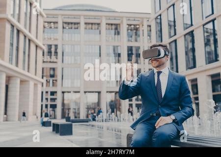 Lächelnder Geschäftsmann mit Stoppeln sitzt auf der Bank auf dem Stadtplatz mit VR-Brille Stockfoto