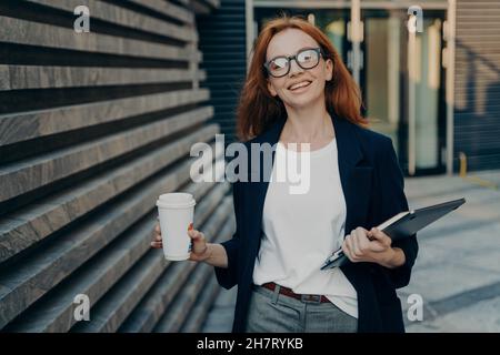 Rotschopf Frau genießt Spaziergang im Freien Getränke zum Mitnehmen Kaffee trägt digitale elektronische Gerät Notizblock Stockfoto