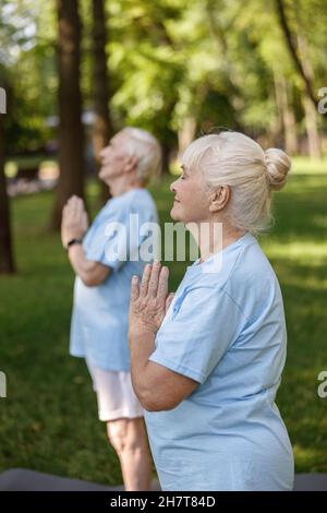 Glückliche ältere Frau hält sich in Namaste die Hände, die mit ihrem Mann im Park Yoga macht Stockfoto
