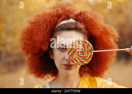 Porträt einer Frau, die im Park in einem gelben Regenmantel mit afro lockigen roten Haaren mit großem Lollipop in der Hand spaziert. Verrückte Ansicht Copy Space Halloween Konzept Stockfoto