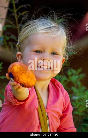 Ein blondes, zweijähriges Mädchen, das ein Osterei hochhält und lächelt. Stockfoto