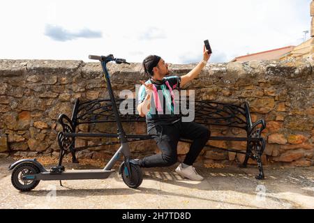 Kaukasischer Mann mit geflochtenem Stirnband und Brille, der ein Selfie mit seinem neuen Elektroroller macht Stockfoto