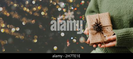 Kleines Geschenk in den Händen der Frau. Nahaufnahme. Geschmückter Weihnachtsbaum auf dem Hintergrund. Helles Weihnachtsbanner mit Platz für Text Stockfoto