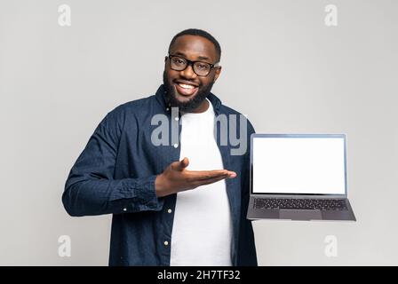 Lächelnder junger afroamerikanischer Kerl in legerer Kleidung, der einen Laptop mit leerem Display auf grauem Hintergrund zeigt. Schwarzer Kerl zeigt mit Handfläche auf leeren Bildschirm, Werbung für neue App, Präsentation von Neuheit Stockfoto