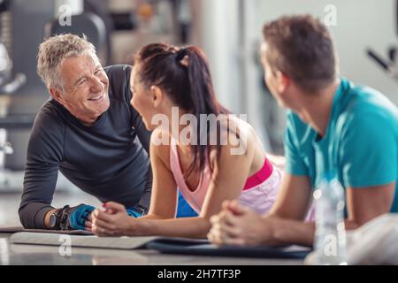 Pensionierter, fittiger Mann lacht zusammen mit jüngeren Fitnessmitgliedern nach dem Training, die auf den Matten auf dem Boden liegen. Stockfoto