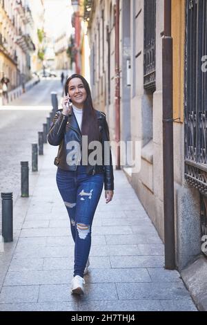 Ganzkörperfrohige junge Frau, die auf dem Smartphone spricht und lächelt, während sie an einem sonnigen Tag auf der Stadtstraße auf dem Bürgersteig läuft Stockfoto