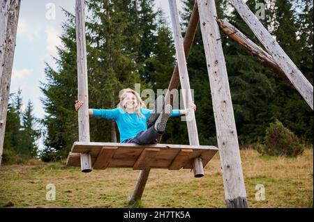 Glückliche Frau, die mit geraden Beinen auf einem Holzsitz sitzt, der an großen Holzstangen befestigt ist und die sie mit ihren Händen bedeckt. Schaukeln auf riesiger Holzschaukel an frischer Luft. Stockfoto
