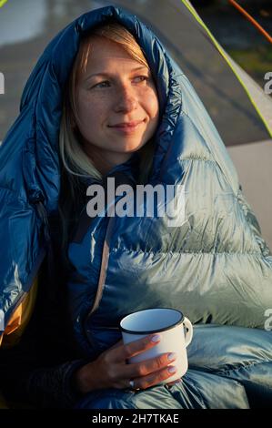 Nahaufnahme einer jungen blonden Frau mit einem zufriedenen Lächeln, umhüllt von einem Schlafsack zum Wandern, mit einem weißen Metallbecher in der Hand und einem Blick nach rechts. Stockfoto