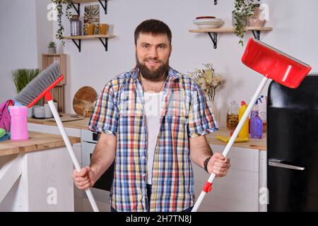 Der junge, gutaussehende bärtige Mann in der Küche, der ein kariertes Hemd trägt, versucht mit einer Kehrschaufel und einem Besen umzugehen. Stockfoto