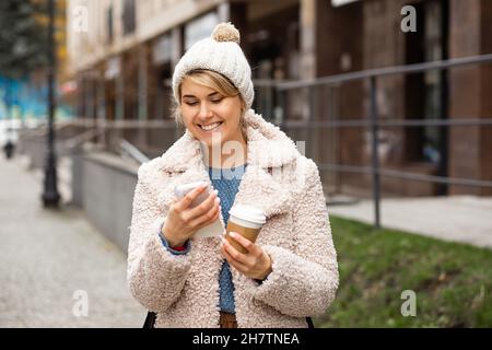 Porträt der jungen hübschen Frau mit Mobiltelefon, SMS-Nachricht, halten Einweg-recycelbare Tasse, heißes Getränk von Kaffee, Tee. Lächelnd, Hut tragend, Stockfoto