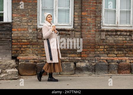 Hübsche junge Frau in beigefarbenem Kunstpelzhut, Mantel mit recycelbarer Einweg-Tasse Kaffee in den Händen geht durch die Stadt im Hintergrund der alten Art roten Bric Stockfoto
