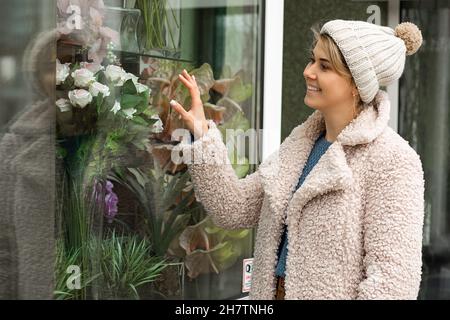 Niedliche junge schöne Frau in gestricktem Hut und beigem Mantel aus Kunstpelz steht am Fenster des Blumenladens, sieht Blumen, genießt, lächelt im Herbst Stockfoto