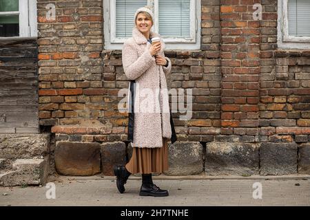 Nette Frau in gestricktem Hut und Pelzmantel aus Kunstpelz mit einer Tasse heißen Kaffee in der Hand geht vor dem Hintergrund eines alten Stalls aus Ziegelsteinen durch die Stadt Stockfoto