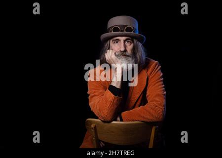 Ein reifer, grauhaariger bärtiger Intellektueller, der einen Steampunk-Hut und eine orangefarbene Jacke trägt, blickt aufmerksam auf die Kamera Stockfoto