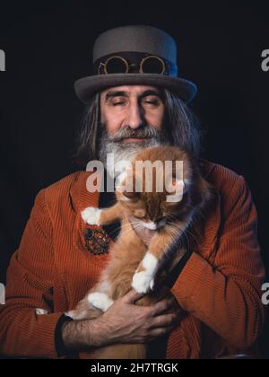 Ein grauhaariger bärtiger Intellektueller im reifen Alter in einem Steampunk-Hut hält eine Ingwerkatze, ein Mann ist glücklich, seine Augen sind geschlossen Stockfoto