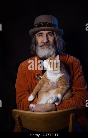 Ein grauhaariger bärtiger Intellektueller im reifen Alter in einem Steampunk-Hut posiert mit einer Ingwerkatze. Stockfoto