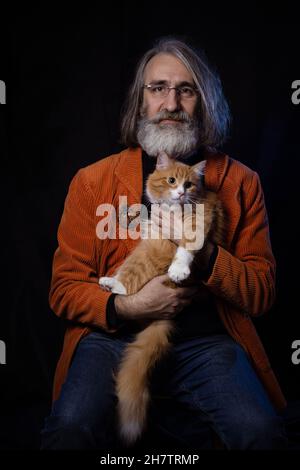 Grauhaariger bärtiger Intellektueller im reifen Alter, der mit einer Ingwer-angepassten Katze posiert Stockfoto