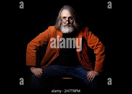 Аn Intellektueller in modischer Brille blickt kritisch auf die Kamera. Ein grauhaariger bärtiger Mann in einer orangefarbenen Jacke. Stockfoto
