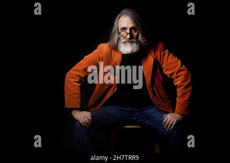 Аn Intellektueller in modischer Brille blickt kritisch auf die Kamera. Ein grauhaariger bärtiger Mann in einer orangefarbenen Jacke. Stockfoto