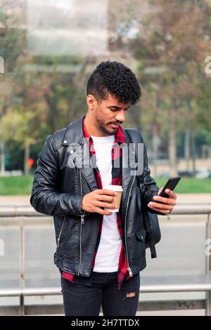 Stilvoller junger lateiner, der sein Telefon an der Bushaltestelle mit einem Kaffee zum Mitnehmen in der Hand, einem Konzept der Technologie und einem urbanen Lebensstil überprüft Stockfoto