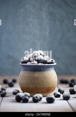 Eine Tasse aus Steinzeug, gefüllt mit Heidelbeeren auf einer keramischen Oberfläche. Staubpulver an die Beeren gesiebt Stockfoto