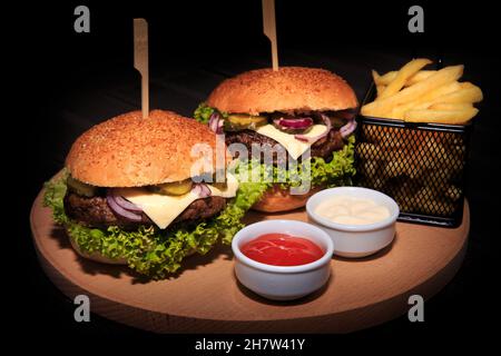 Klassische Cheeseburger mit Rinderpastete, Gurken, Käse, Tomaten, Zwiebeln, Salat, Ketchup und Senf auf einer Holzplatte isoliert auf schwarzem Hintergrund. Stockfoto