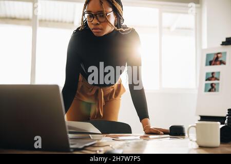 Kreative junge Fotografin, die in ihrem Heimbüro arbeitet. Junge Freiberuflerin, die an einem Schreibtisch mit Fotoausrüstung steht. Selbstständige Frau uns Stockfoto