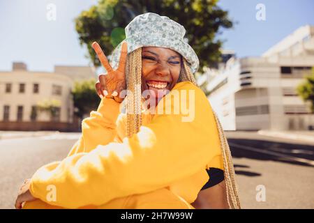 Sorglose junge Frau, die an einem Sommertag im Freien das Friedenszeichen macht. Glückliche junge Frau, die Spaß hat und fröhlich die Kamera anlächelt Stockfoto