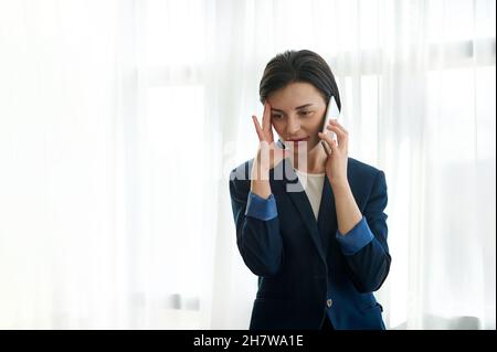 Attraktive Brünette, weibliche Angestellte, müde Geschäftsfrau, die Hand auf ihren Tempel legt, während sie ein langweiliges Gespräch am Telefon führt Stockfoto