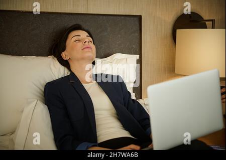 Schöne, elegante kaukasische junge Geschäftsfrau mit Laptop, die mit geschlossenen Augen auf dem Bett liegt und sich nach einem harten Arbeitstag erschöpft fühlt Stockfoto