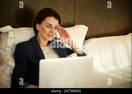 Müde, aber glücklich junge Freiberufler, Unternehmerin, Geschäftsfrau arbeitet auf einem Laptop, während auf dem Bett liegend, lächelnd niedlich freut sich über eine erfolgreiche und p Stockfoto