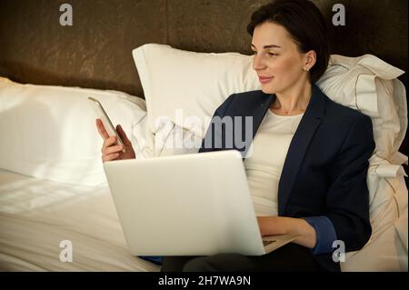 Junge kaukasische Geschäftsfrau, die Mobiltelefon und Laptop benutzte, während sie sich auf dem Bett lag und sich nach einer Geschäftsreise in einem Hotelzimmer entspannen konnte Stockfoto