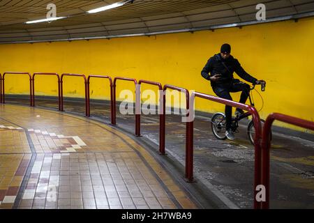 Ein schwarzer Mann fährt mit seinem Fahrrad durch einen Tunnel in Venedig Mestre mit einer gelb gefliesten Wand neben roten Metallgeländern und schaut auf sein Smartphone. Stockfoto