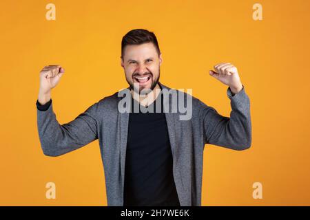 Verkürztes Porträt eines glücklichen und aufgeregten Mannes in grauer Strickjacke, der fröhlich isoliert auf gelbem Hintergrund posiert. Gewinnen Stockfoto
