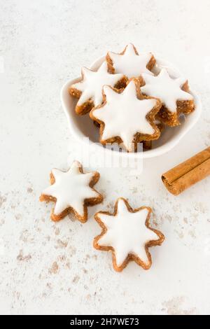 Zimtstern-Kekse, traditionelle deutsche Zimtsterne in einer weißen Schale auf gemustertem Hintergrund mit Zimtstange. Speicherplatz kopieren. Stockfoto