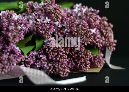 Ein Bouquet von lila Gartenfliedern in handwerklicher Verpackung mit einem rosa Band auf dunklem Hintergrund. Grußkarte. Selektiver Fokus. Horizontales Foto. Stockfoto