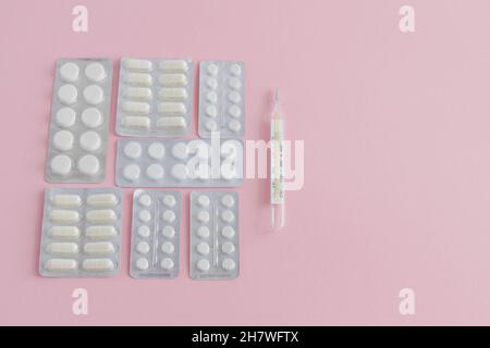 Quecksilberthermometer zur Messung der Körpertemperatur in einem Set mit Tabletten und Kapseln in Blasen zur Behandlung auf einem rosa Hintergrund Stockfoto