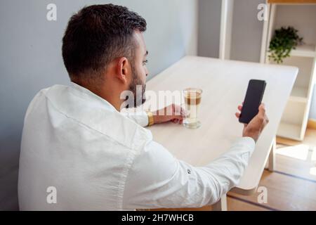 Mann mit einem Handy. Mann, der auf das Handy schaut. Mann im Café mit Handy. Mann, der Kaffee im Café hat. Stockfoto