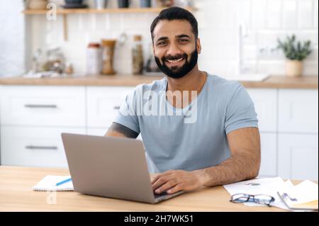 Porträt eines positiv freundlichen bärtigen indischen Mannes, der zu Hause am Tisch in der Küche sitzt, fernab von zu Hause mit einem Laptop arbeitet, direkt auf die Kamera blickt und lächelt Stockfoto