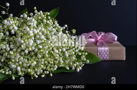 Studio-Foto. Grußkarte. Ein Bouquet von duftenden weißen Lilien des Tals auf dunklem Hintergrund mit einem Geschenk in handgefertiger Verpackung mit einem rosa Band. Stockfoto