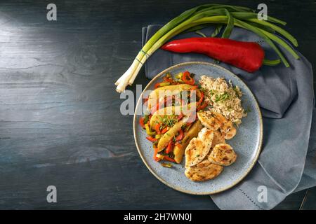 Gebratene Hähnchenscheiben mit braunem Vollkornreis und Gemüse wie Paprika, Lauch und Birne auf einem blauen Teller und einem dunklen Holztisch, Platz kopieren, t Stockfoto