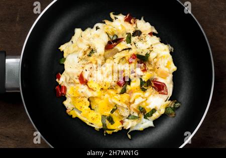 Rühreier auf einer schwarzen Pfanne über dunkler Holzplatte, gekocht mit Zwiebeln, roten Chilischoten, Paprikeblättern, flach liegend, Draufsicht Stockfoto