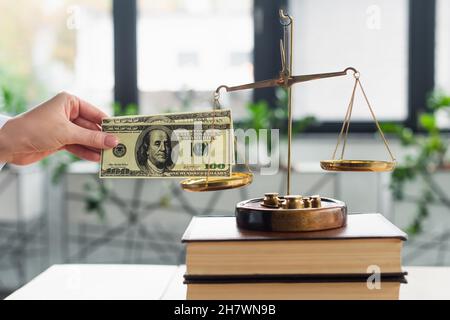 KIEW, UKRAINE - 22. SEPTEMBER 2021: Beschnittene Ansicht der Frau, die Dollar in der Nähe von Justizwaagen auf Büchern im Amt hält, Antikorruptionskonzept Stockfoto