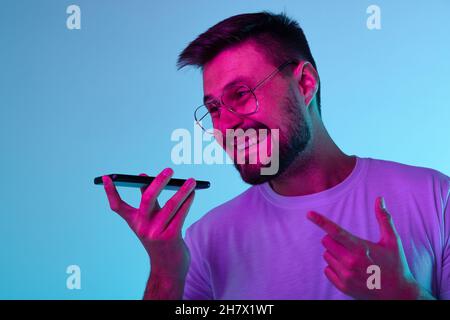 Beschnittenes Porträt eines aufgeregten Mannes, der fröhlich Sprachnachrichten aufzeichnete, wobei das Telefon auf blauem Hintergrund in Neonlicht isoliert war Stockfoto