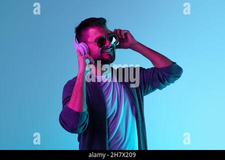 Beschnittenes Porträt eines Mannes in einer Sonnenbrille, der fröhlich Musik in weißen Kopfhörern hört, die auf blauem Hintergrund isoliert sind Stockfoto