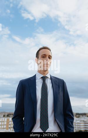 Ein junger Geschäftsmann in Anzug und Krawatte, der ernsthaft auf die Kamera schaut, die draußen auf einem Dach mit bewölktem Himmel steht Stockfoto