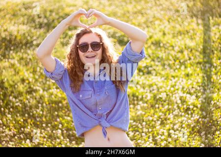 Junges blondes Landmädchen in Denim-Hemd mit Knöpfen, das heiße jean-Shorts trägt, posiert im Sommer zur goldenen Stunde im Freien und schaut verführerisch auf die Kamera Stockfoto